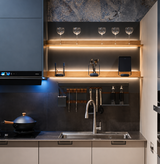 Modern kitchen interior with a dark gray backsplash, built-in wood floating shelves with under-shelf lighting, wine glasses, glassware, bottles, and a stainless steel sink with a curved faucet. A black range hood is visible on the left, and a blue-lit cookware pot sits on the stovetop.