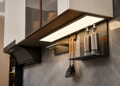 Modern wall-mounted kitchen cabinetry and lighting in a real kitchen setting, with a black range hood, white upper cabinets, under-cabinet lighting, wall shelves, stainless steel bottles, and hanging utensils against a gray stone backsplash.