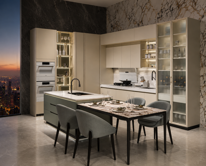 A modern L-shaped kitchen with cream cabinets, marble countertops and island, built-in ovens, glass-front storage cabinets, and a dining table with four gray chairs. The kitchen is set in a high-rise apartment with a city skyline and sunset visible through a large window on the left.