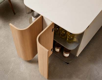 A modern kitchen cabinet with a white top and light wood curved doors, shown open to reveal shelves with bottles and jars inside, set on a gray concrete floor.