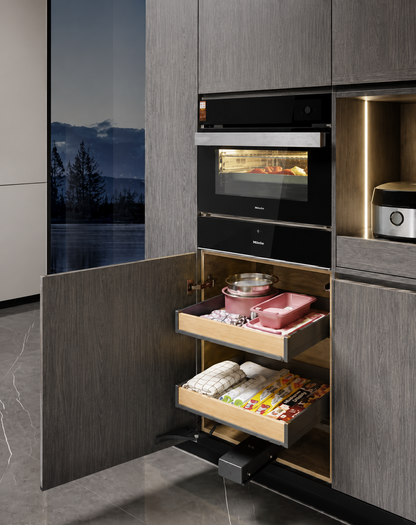 A modern kitchen cabinet with dark wood grain doors, featuring a built-in black oven and two open pull-out drawers stocked with cookware and packaged food. A large window on the left shows an outdoor mountain landscape at dusk.