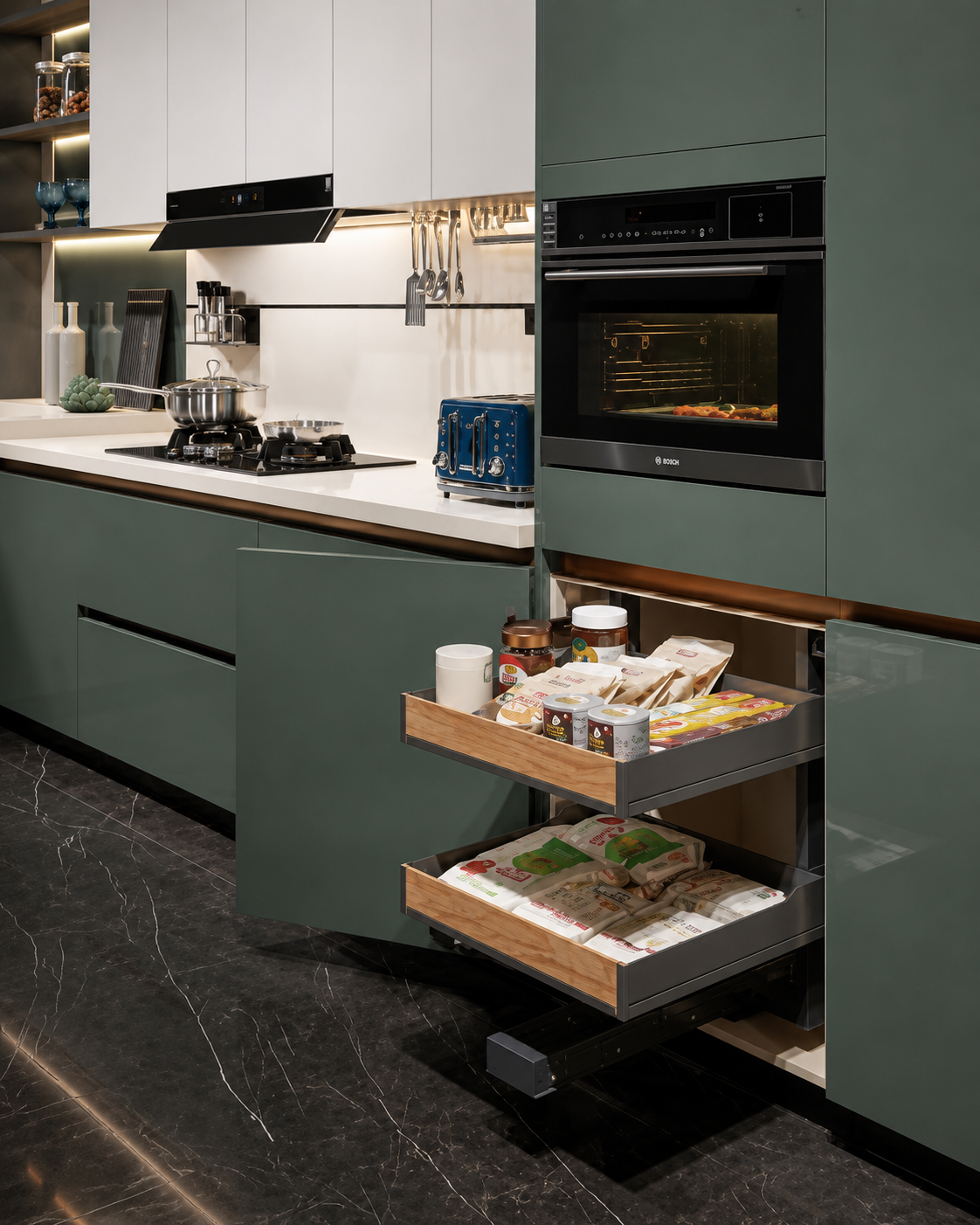 A modern kitchen interior with sage green cabinets, a built-in black oven, white countertops, and two open pull-out drawers showing packaged food items. The floor is dark marble-look tile.