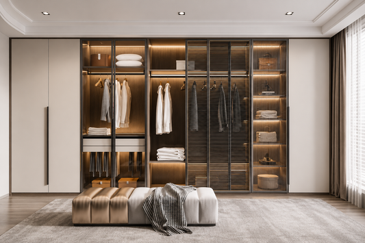 A modern walk-in closet system with glass doors, warm interior lighting, hanging garments, folded textiles, and a matching bench in a styled bedroom or dressing room setting.