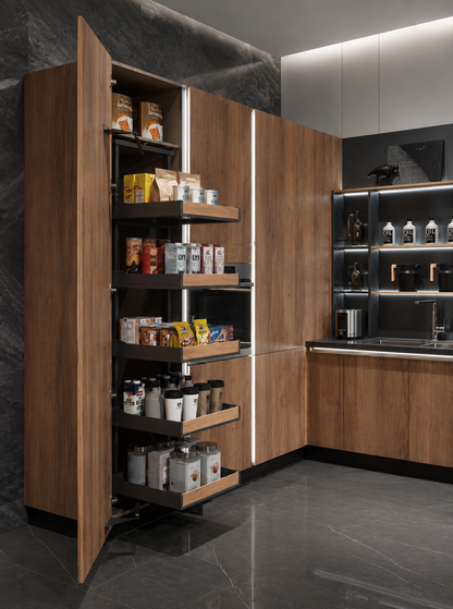 A modern kitchen interior with a tall wooden pantry cabinet on the left, shown open to reveal multiple pull-out shelves stocked with jars, cans, and packaged goods. The kitchen has dark gray stone flooring and backsplash, wood cabinetry, and built-in under-cabinet lighting.