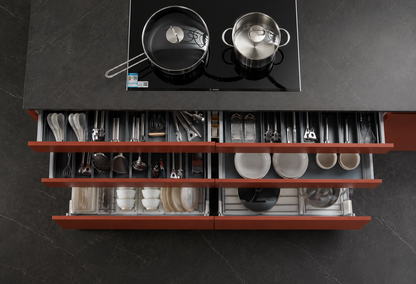 An overhead view of a modern kitchen cabinet with two open lower drawers organized with cookware and dishes, set beneath a black induction cooktop on a dark stone floor.