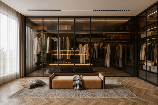 A modern walk-in closet with black-framed glass doors, warm lighting, hanging garments, and a tan upholstered bench placed on a gray rug in front.
