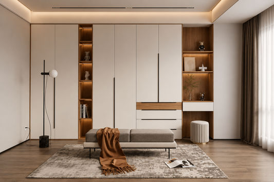 A modern built-in wardrobe and storage system in a minimalist bedroom. The system features large off-white doors with slim vertical handles, warm wood shelving with integrated LED lighting, a matching desk unit, and a small gray bench with a brown throw blanket on a textured rug.