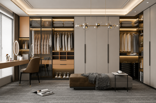 A modern walk-in closet room with tall light gray wardrobe doors, open shelving with hanging garments, a vanity desk with a chair, a bench with a throw blanket, and a small side table. The space is styled in warm neutral tones with wood accents, recessed lighting, and a chandelier.