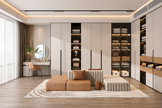 A modern home office or dressing room interior featuring a large built-in wardrobe/closet system with cream and black doors, shelving, and lighting. In the foreground is a low tan ottoman-style seating piece with ribbed cream cushions, a matching rectangular bench, and a round brown accent, all arranged on a light rug over wood flooring.