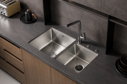 A modern double-basin stainless steel sink with a chrome faucet installed in a dark gray countertop with light wood cabinets.