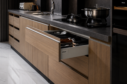 A modern kitchen cabinetry setup with light brown wood-grain lower cabinets, a dark gray countertop, and an open drawer showing kitchen utensils. A built-in black appliance is visible on the right side.